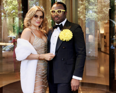 A stylish couple in formal attire posing at a hotel entrance: woman in a beaded sheer gown with a white fur wrap, man in a black tuxedo with a yellow flower on his lapel and yellow glasses.