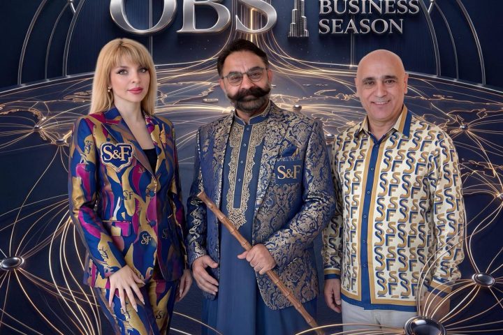 Three people pose in front of a CBS One Business Season backdrop; a woman in a colorful patterned suit stands on the left, a man in an ornate blue jacket center holds a staff, and another man in a patterned shirt stands on the right.