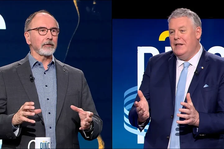 Two men in business suits speak in a split-screen TV interview, gesturing with their hands.