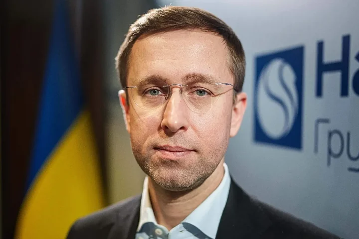 Close-up of a man in glasses in a suit speaking at a press event, with a blue-yellow flag in the background.