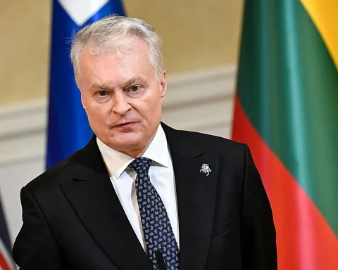 Older man in a black suit and patterned tie, indoors, with colorful flags in the background.