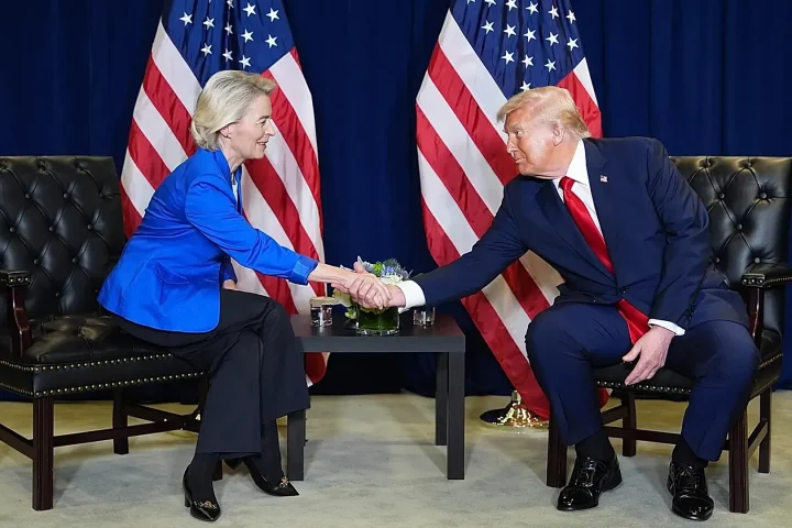 Two people in formal attire shake hands across a small table, with American flags in the background.",