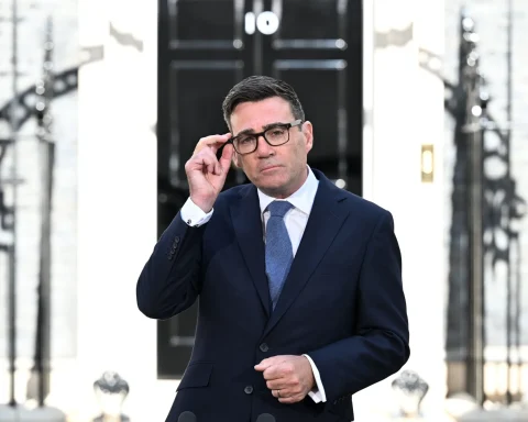 Man in a navy suit and glasses adjusting his frame outside a building entrance marked with the number 10.