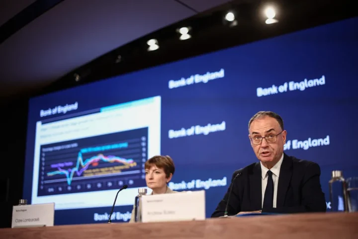 Two Bank of England officials speak at a press conference, with a large screen showing a multi-line chart in the background.
