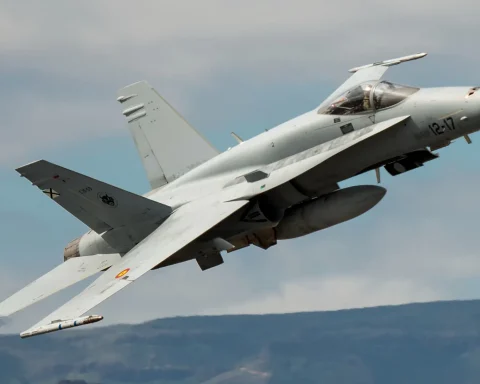 Gray military fighter jet banking during flight, cockpit canopy visible, Spanish Air Force markings on wing, mountainous landscape below