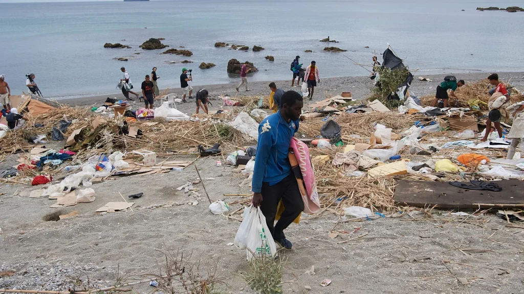 Volunteers cleaning a littered beach, collecting trash into bags as debris covers the shoreline with calm water in the background.
