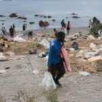 Volunteers cleaning a littered beach, collecting trash into bags as debris covers the shoreline with calm water in the background.