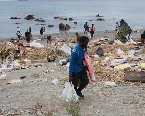 Volunteers cleaning a littered beach, collecting trash into bags as debris covers the shoreline with calm water in the background.