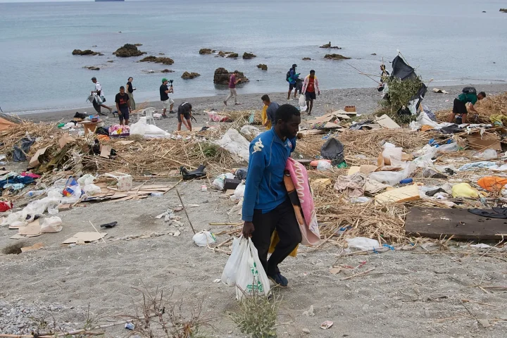 Volunteers cleaning a littered beach, collecting trash into bags as debris covers the shoreline with calm water in the background.
