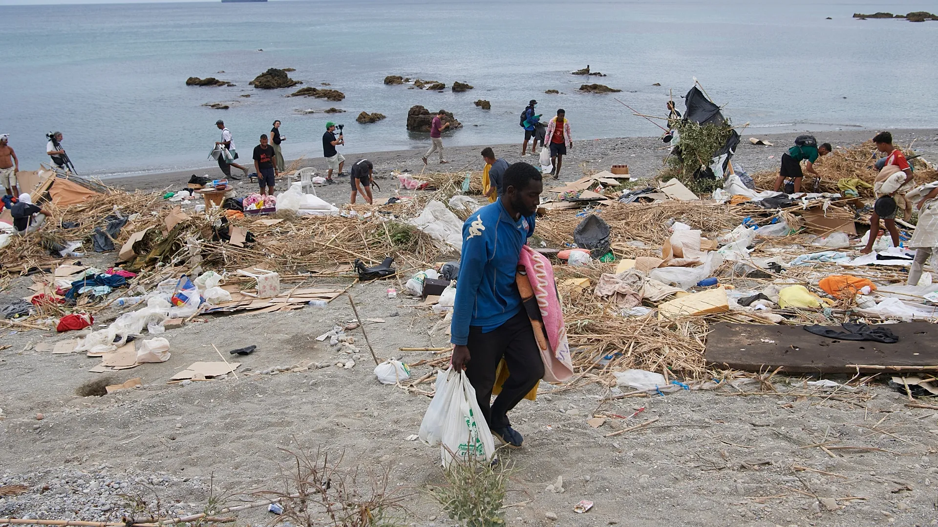 Volunteers cleaning a littered beach, collecting trash into bags as debris covers the shoreline with calm water in the background.