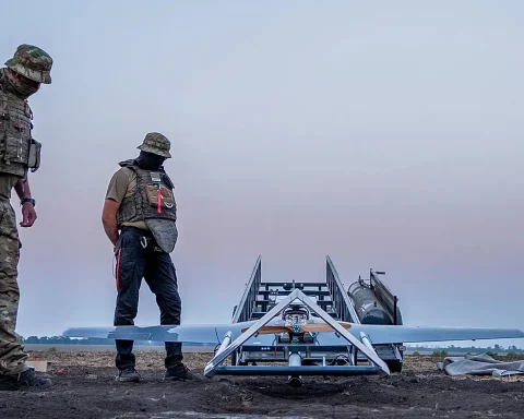 Two soldiers in camouflage gear stand on a dirt field beside a wheeled launch rig carrying a small aircraft, under a dusky sky.