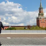 Man in a dark suit walks across a cobblestone plaza with the Moscow Kremlin towers in the background under a partly cloudy sky.
