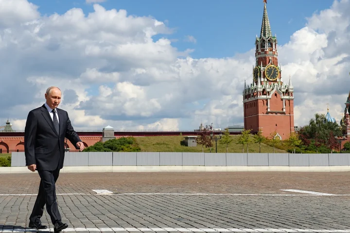 Man in a dark suit walks across a cobblestone plaza with the Moscow Kremlin towers in the background under a partly cloudy sky.