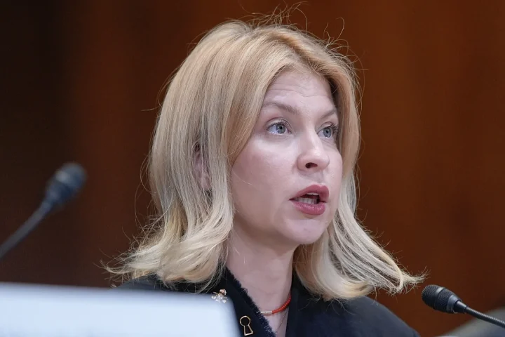 Blonde woman speaking into a desk microphone at a panel, with a laptop visible in the foreground.