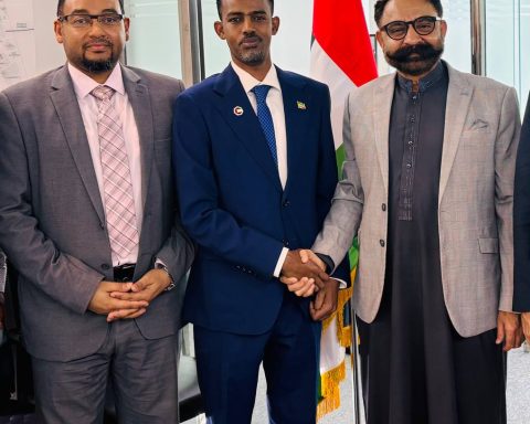 Three men in suits shake hands in a formal indoor setting with a flag in the background, indicating a business or diplomatic meeting.
