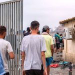 Civilians wait in a line behind a tall metal gate at a security checkpoint, with a soldier standing guard nearby.